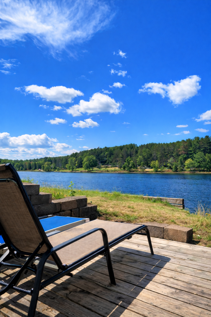 This image captures a tranquil lakeside scene with a reclining lounge chair in the foreground, offering a peaceful view of the calm waters. The chair sits on a wooden deck, positioned for optimal relaxation under the bright blue sky, dotted with fluffy white clouds. The lush green landscape stretches across the horizon, with trees lining the opposite shore, creating a serene and picturesque atmosphere. The perfect spot to unwind and enjoy the natural beauty.