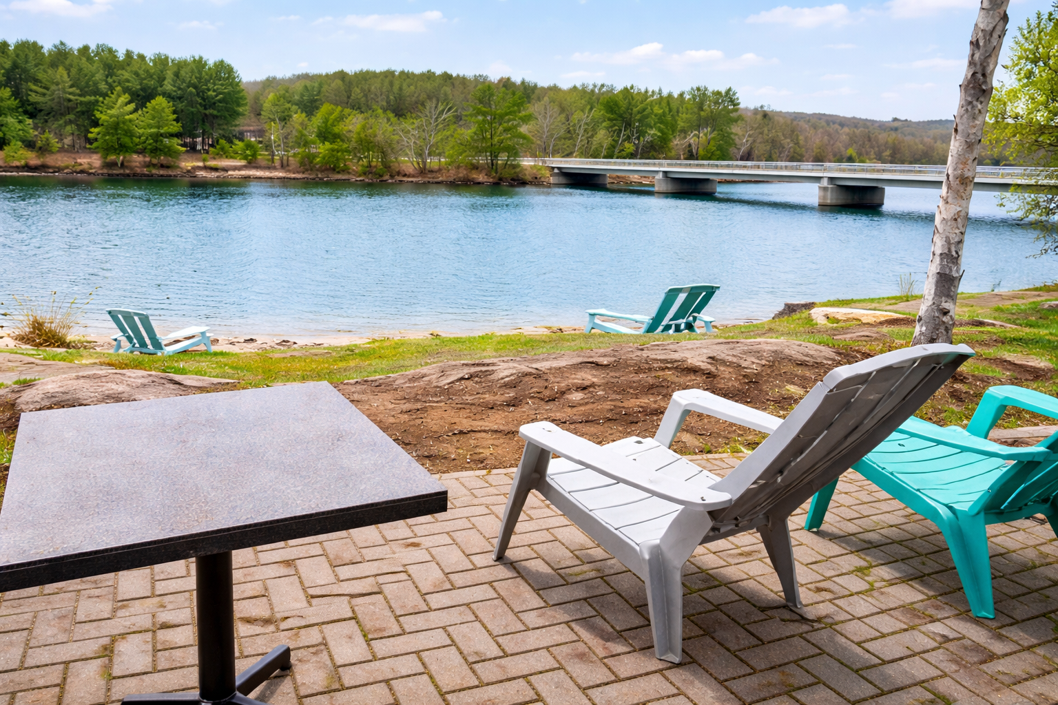 This image captures a peaceful lakeside patio with a scenic view. A cozy, paved seating area features a dark table and a mix of white and turquoise Adirondack chairs, all positioned to overlook a serene lake. In the distance, a bridge stretches across the water, framed by lush greenery on either side. The bright, natural lighting enhances the tranquil atmosphere, offering a perfect spot for relaxation by the water